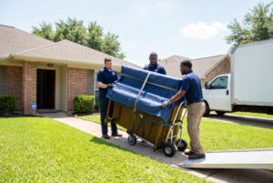 Leo's Moving Services professional piano movers rolling a wrapped upright piano toward a moving truck outside a Houston home